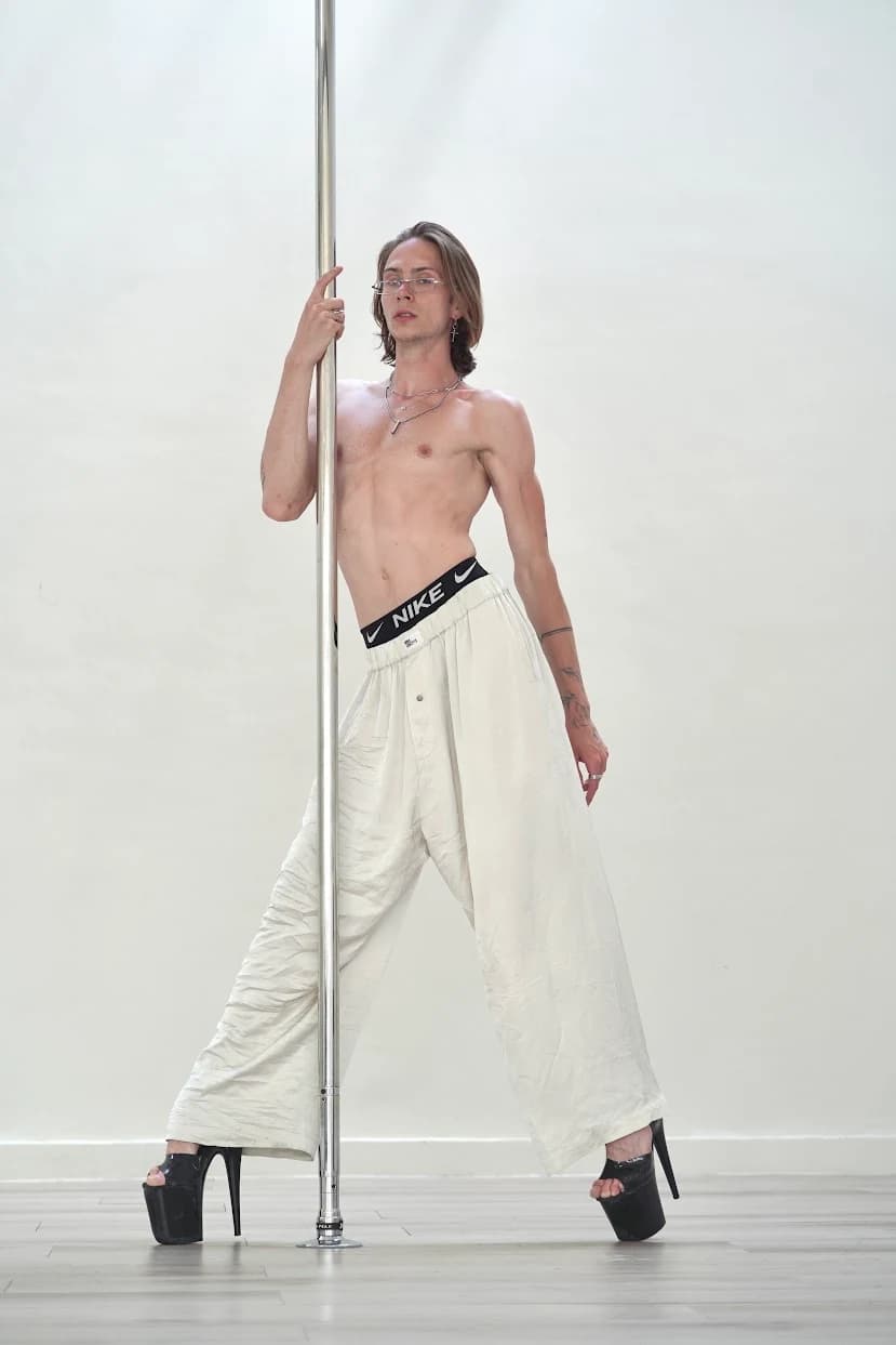 Ilja Med beside a pole in a light studio, wearing white trousers and black heels